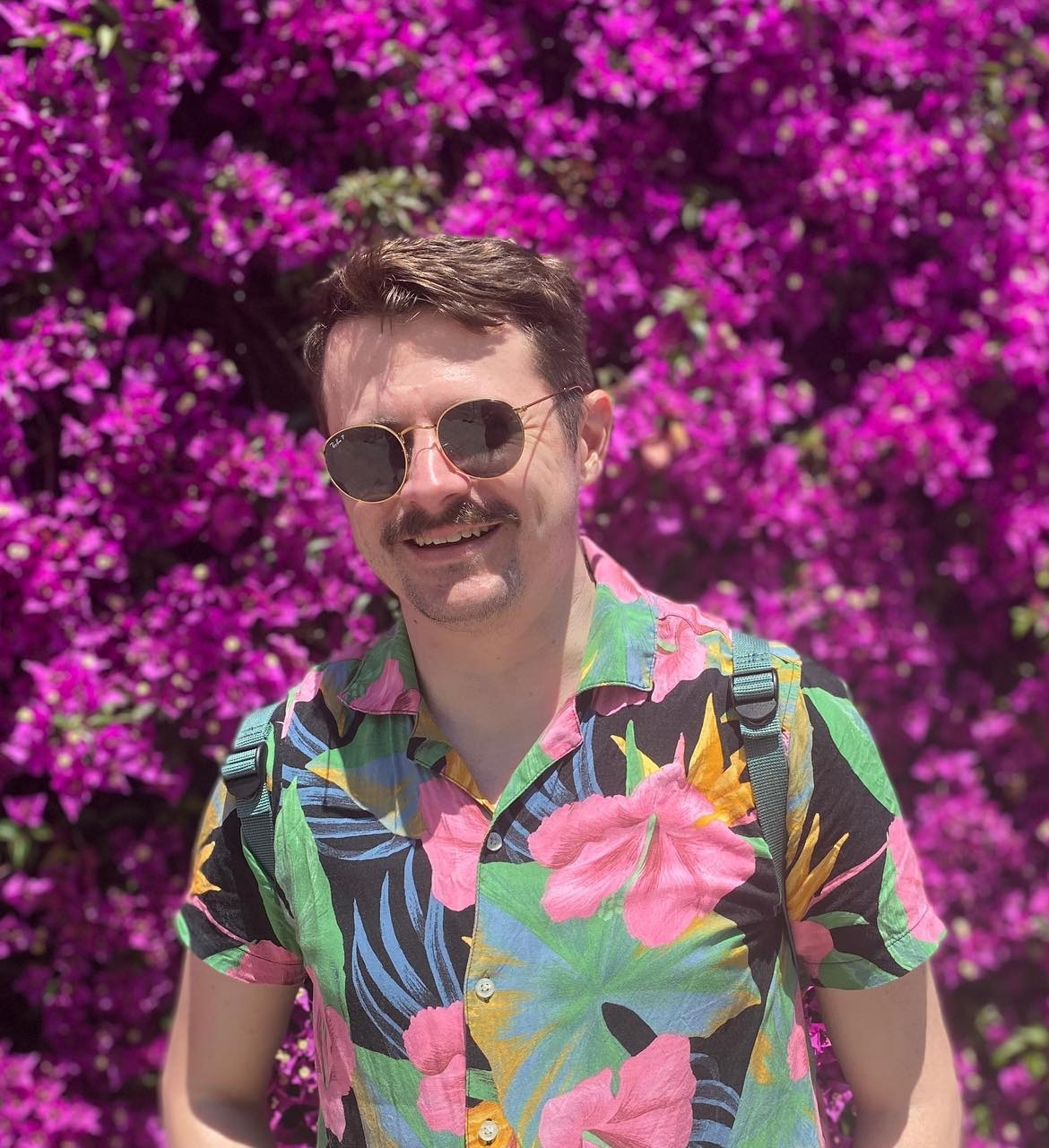 Brendan Miller smiling in front of bright pink bougainvillea, wearing a tropical shirt and aviator sunglasses